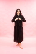 A woman stands barefoot against a solid pink studio backdrop, wearing a long black skirt with a matching cropped button-front jacket.