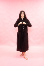 A woman stands barefoot against a solid pink studio backdrop, wearing a long black skirt with a matching cropped button-front jacket.