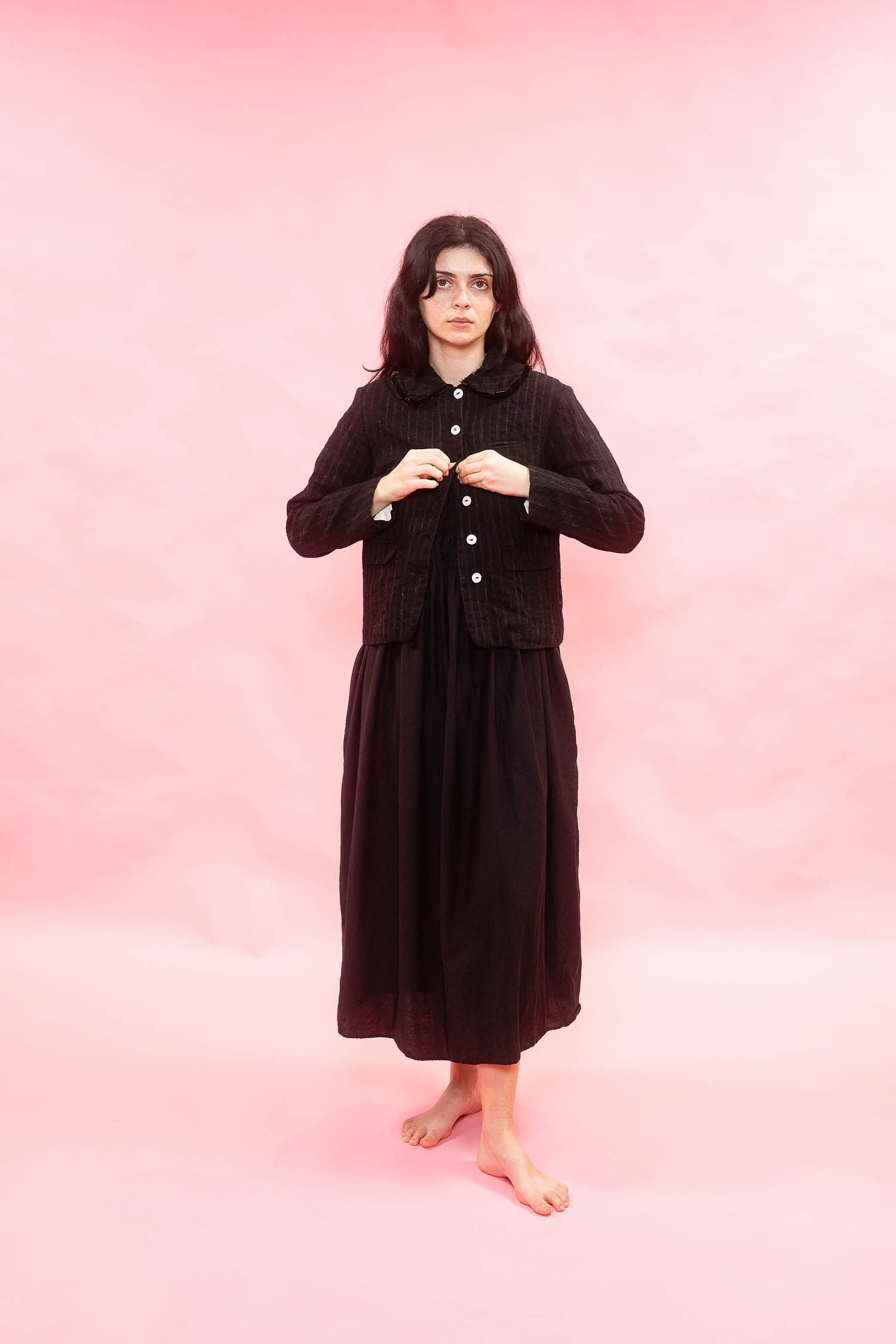 A woman stands barefoot against a solid pink studio backdrop, wearing a long black skirt with a matching cropped button-front jacket.