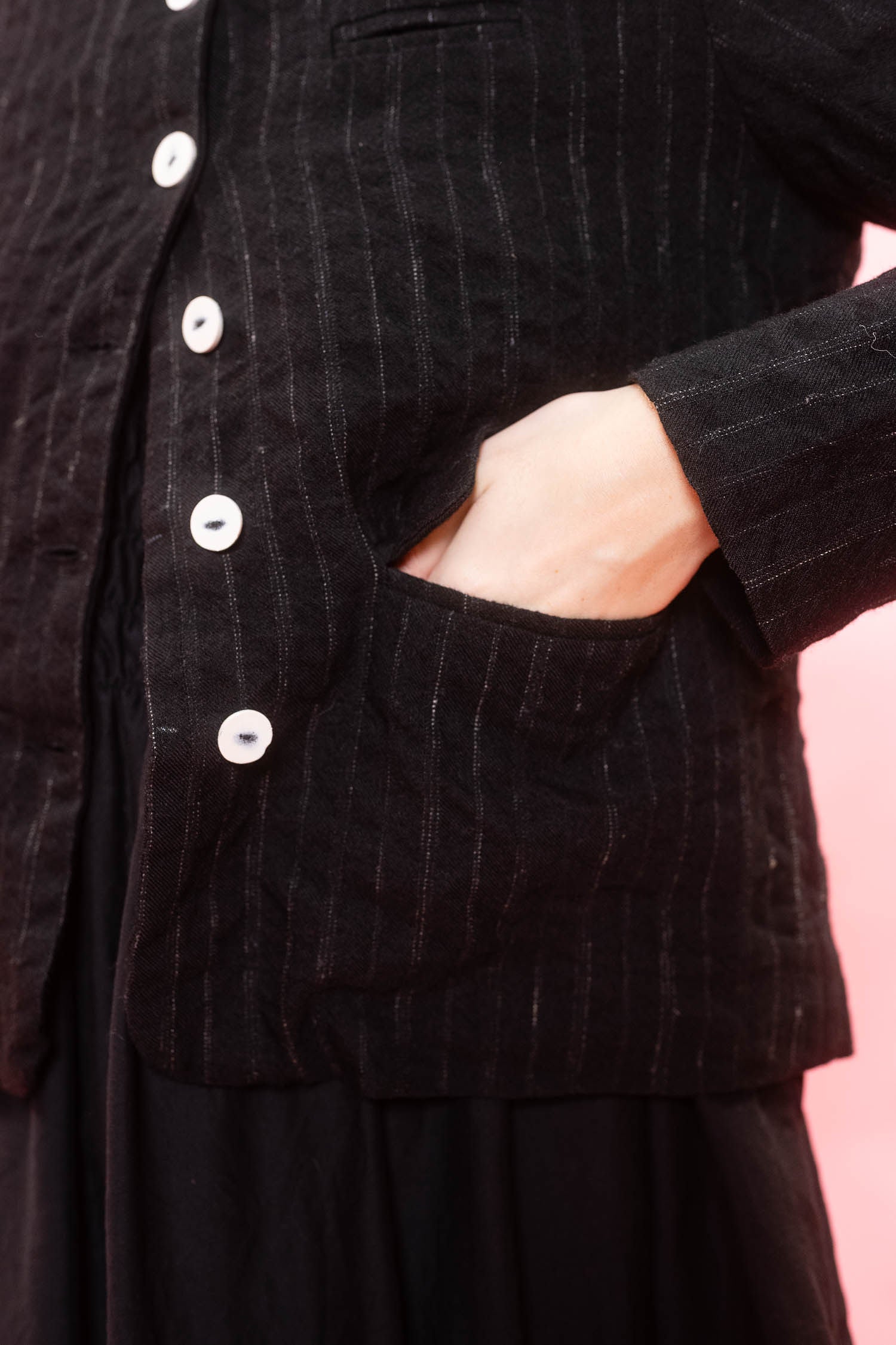 Close-up of a black textured jacket with thin vertical pinstripes, white buttons, and a side pocket with a hand inserted, set against a solid pink background.