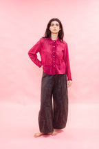 A woman stands barefoot against a solid pink studio backdrop, wearing a bright pink long-sleeve button-front shirt and dark charcoal wide-leg pants.