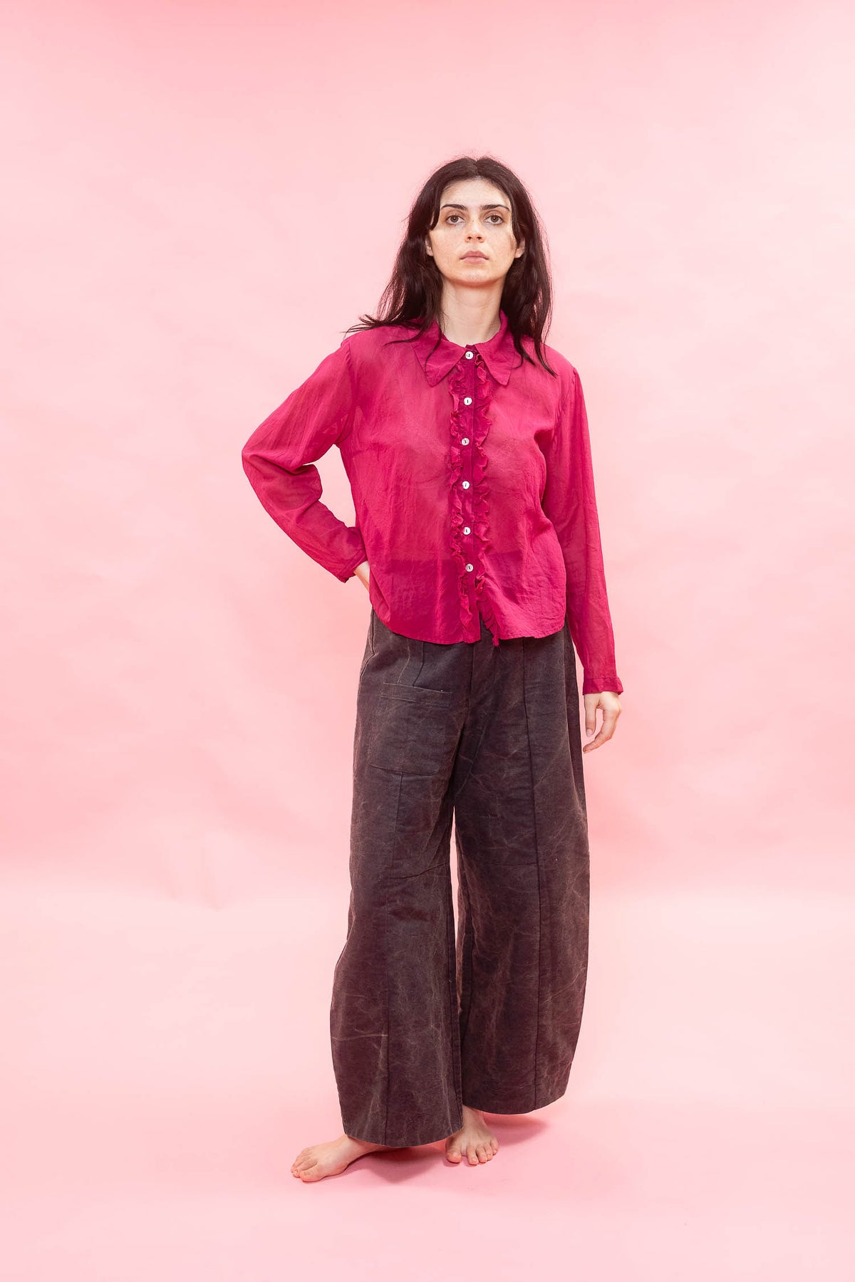 A woman stands barefoot against a solid pink studio backdrop, wearing a bright pink long-sleeve button-front shirt and dark charcoal wide-leg pants.