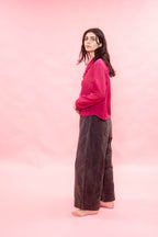 A woman stands barefoot in profile against a solid pink studio backdrop, wearing a bright pink long-sleeve button-up shirt and dark brown wide-leg pants.