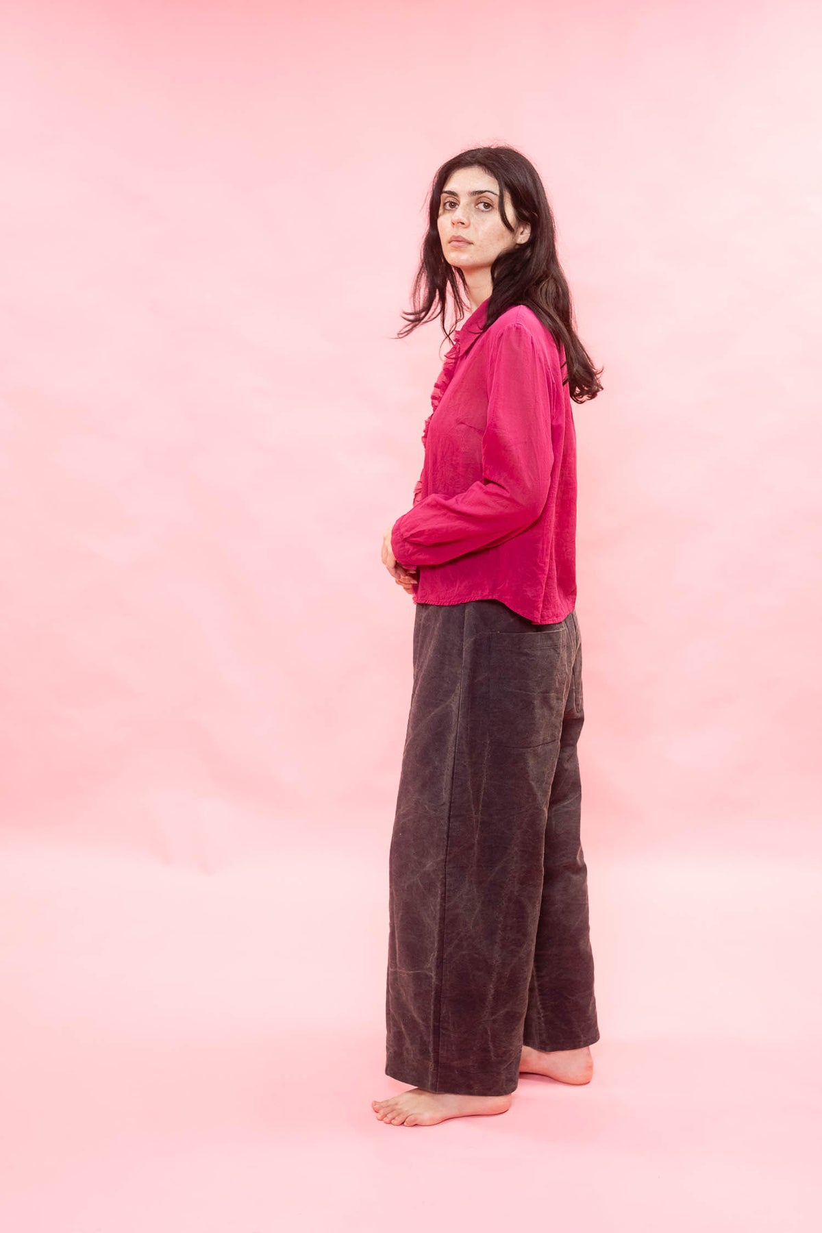 A woman stands barefoot in profile against a solid pink studio backdrop, wearing a bright pink long-sleeve button-up shirt and dark brown wide-leg pants.