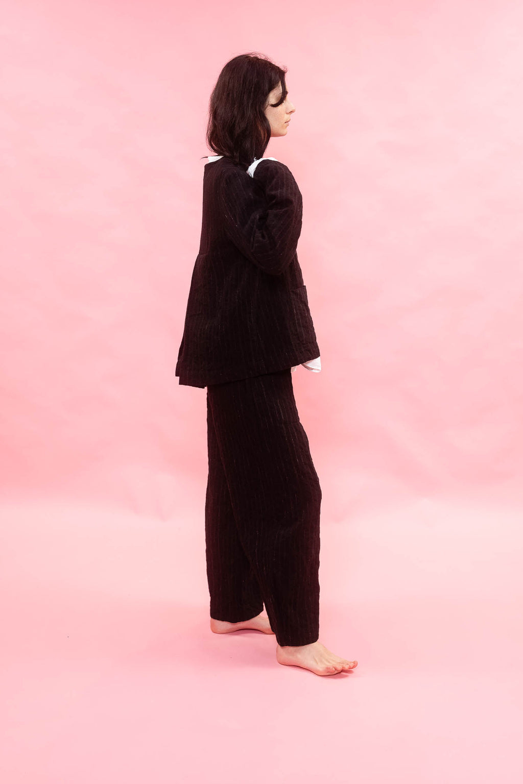A woman stands barefoot in profile against a solid pink studio background, wearing a black wool pinstripe jacket and matching wide-leg trousers.