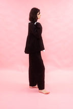 A woman stands barefoot in profile against a solid pink studio background, wearing a black wool pinstripe jacket and matching wide-leg trousers.