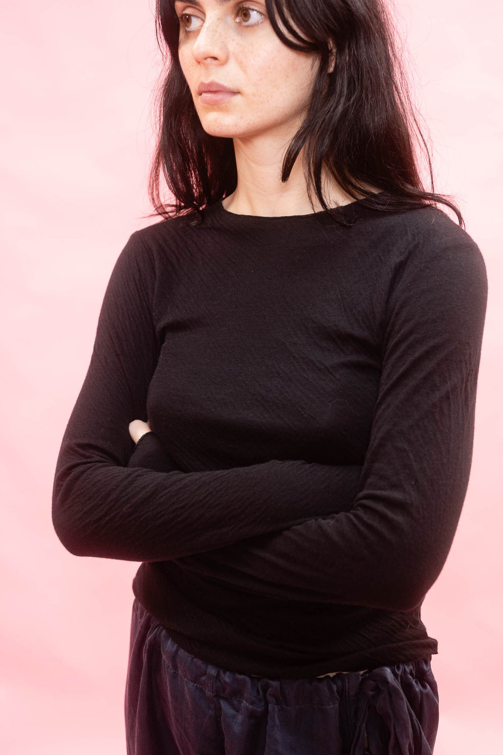 A woman with dark hair wearing a black long-sleeve crewneck tee, cut from Japanese cotton double jersey, standing with arms crossed against a solid pink background.
