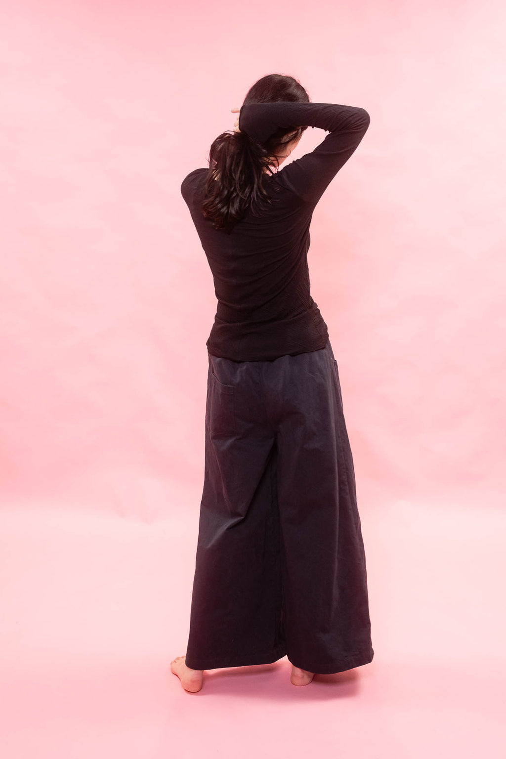 A woman viewed from behind wearing a black long-sleeve top and very wide-leg navy pants, standing barefoot against a solid pink studio background.