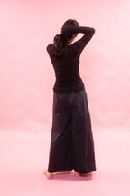 A woman viewed from behind wearing a black long-sleeve top and very wide-leg navy pants, standing barefoot against a solid pink studio background.
