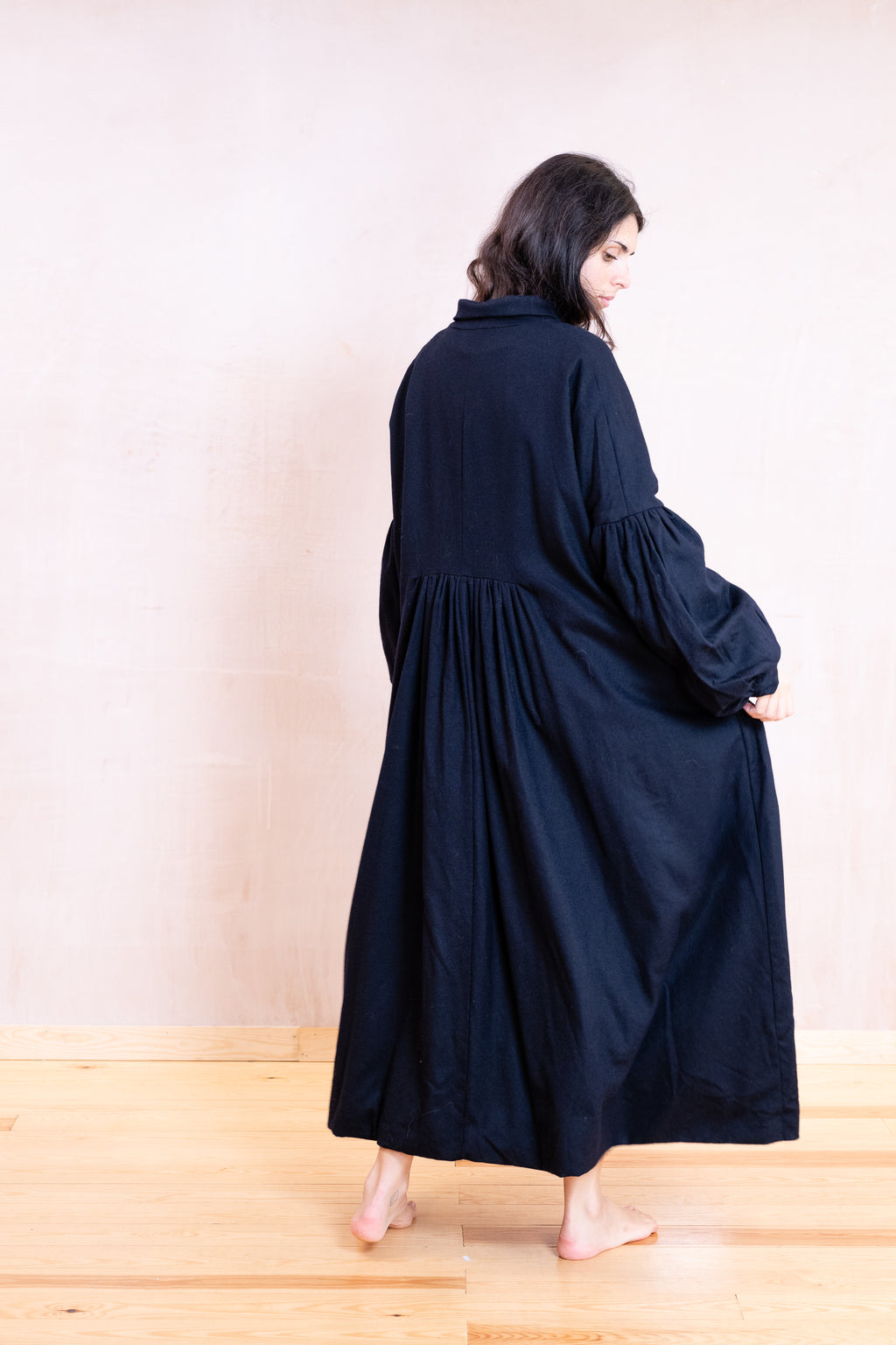 A woman wearing a long, loose-fitting black maxi dress with voluminous balloon sleeves, shown from the back in a minimalist room with a light beige wall and wooden floor.