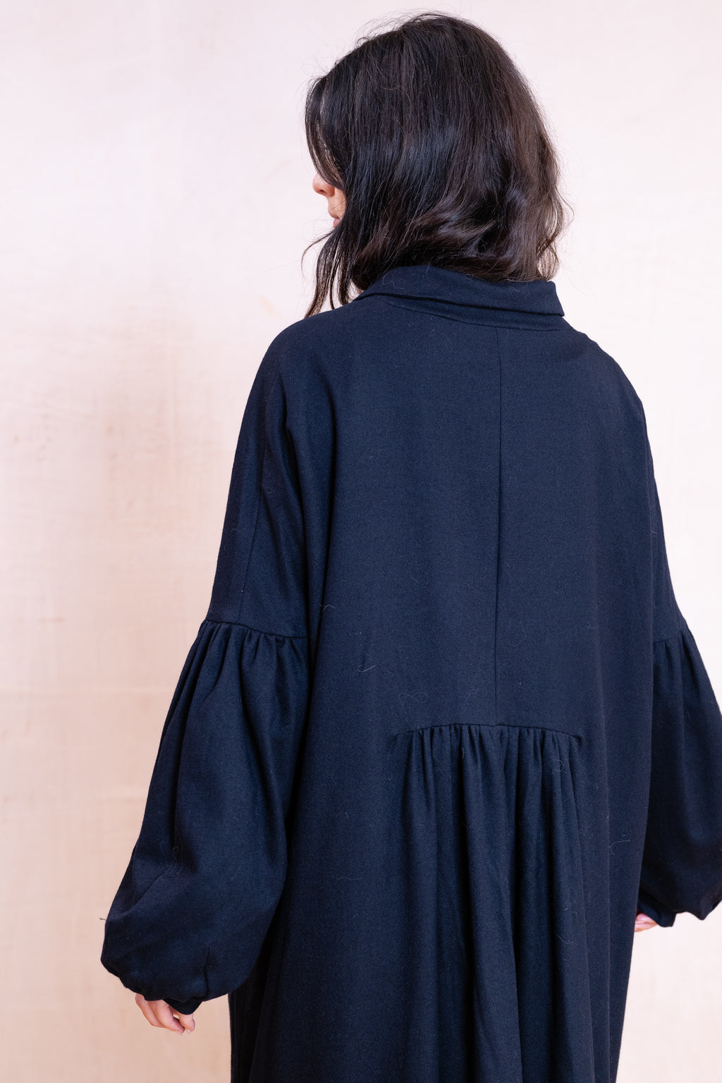 A person shown from the back wearing a loose, dark navy long-sleeve dress with gathered balloon sleeves and a gathered waist seam, set against a plain light beige wall.