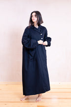 A woman stands barefoot in a studio wearing a long black robe with wide sleeves and front buttons, against a plain light beige wall and wooden floor.