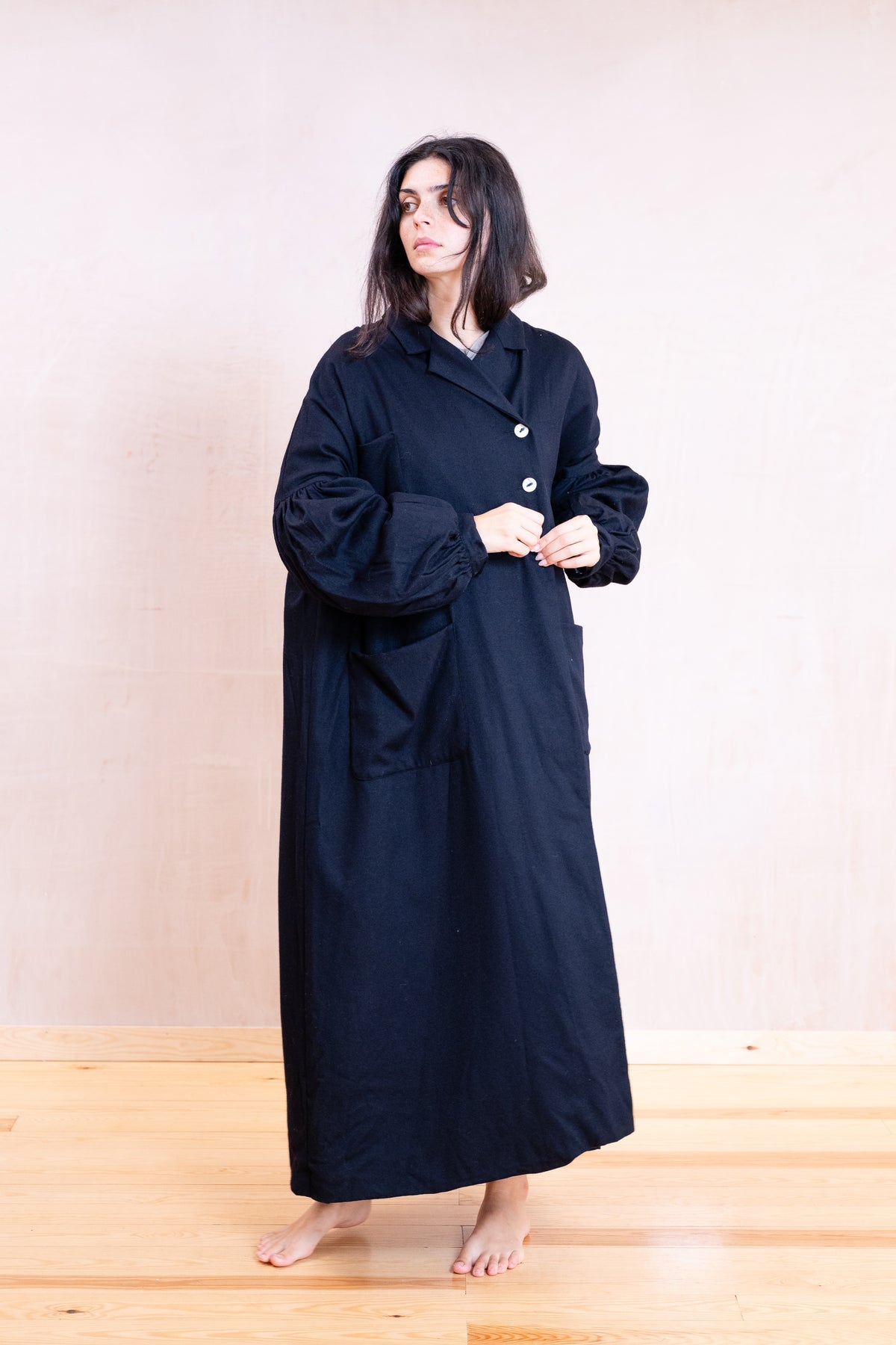 A woman stands barefoot in a studio wearing a long black robe with wide sleeves and front buttons, against a plain light beige wall and wooden floor.