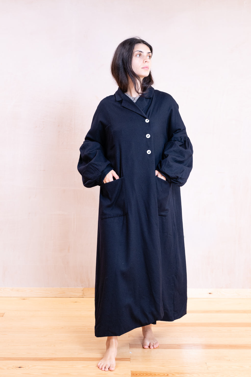 A woman stands barefoot in a studio wearing a long, loose-fitting dark coat with large front pockets and a buttoned collar. The coat appears very dark navy or near-black, with a light neutral background and light wood floor.