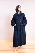 A woman stands barefoot in a studio wearing a long, loose-fitting dark coat with large front pockets and a buttoned collar. The coat appears very dark navy or near-black, with a light neutral background and light wood floor.