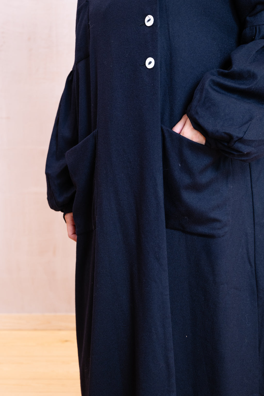 A close-up of a person wearing a long, loose-fitting dark navy coat with large front pockets and white buttons, standing indoors against a light beige wall and pale wood floor.