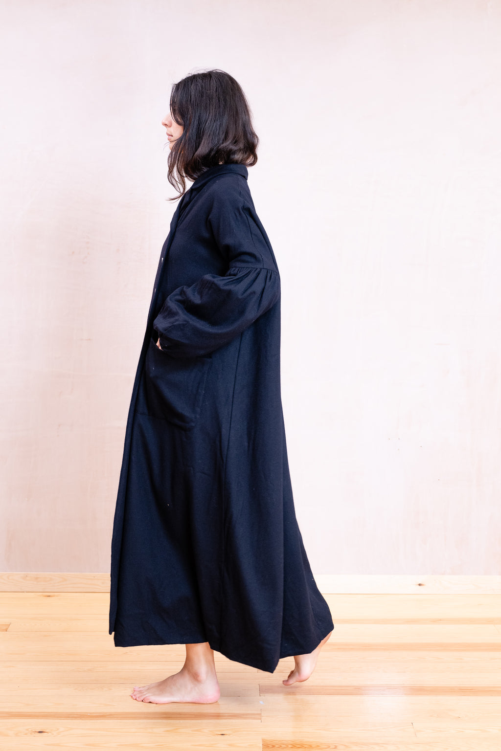 A woman wearing a long, loose-fitting black coat or robe stands barefoot on a light wood floor against a plain beige wall, shown in side profile.