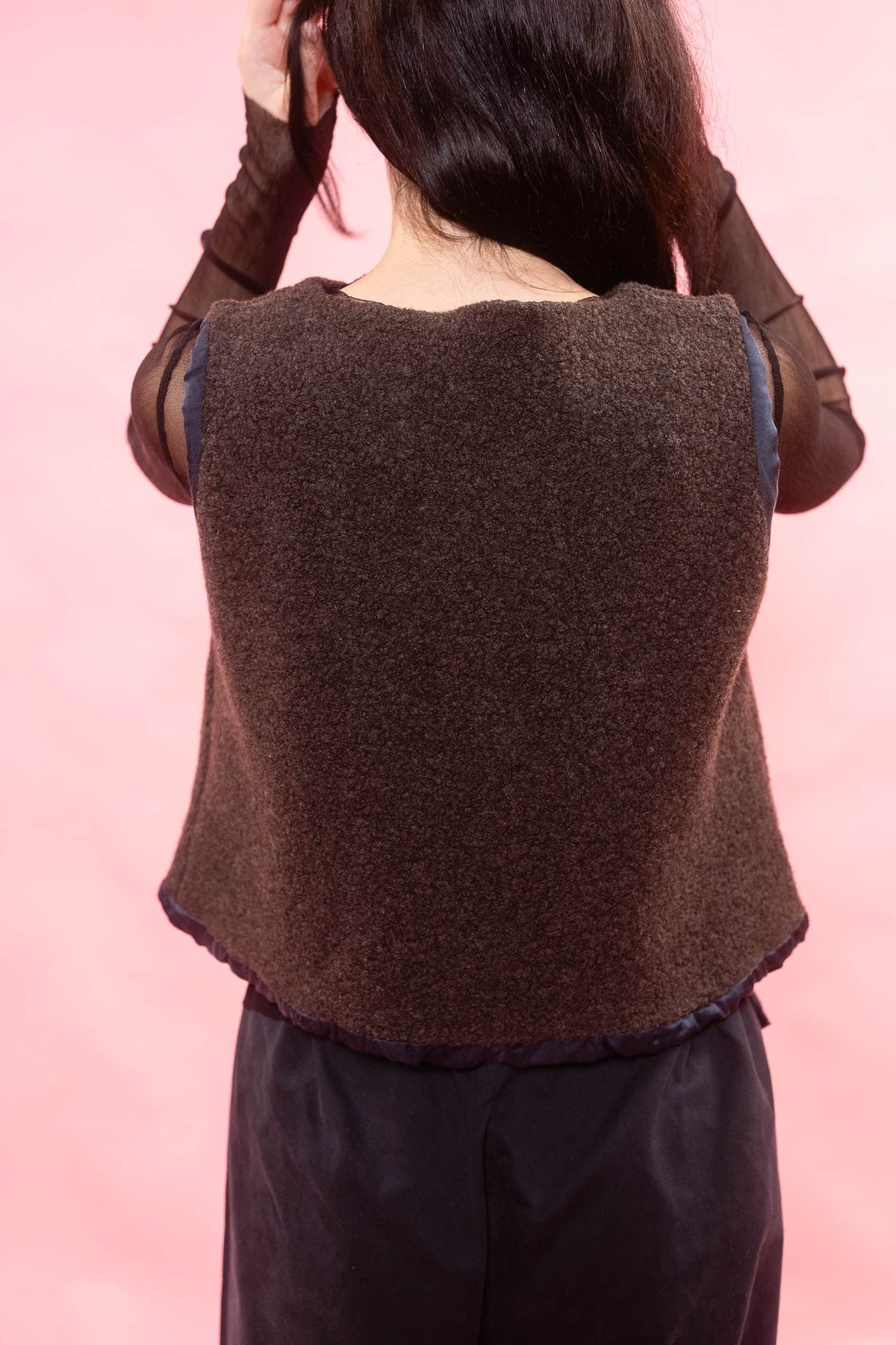 A person shown from the back wearing a brown textured knit sleeveless top or vest over sheer dark brown sleeves, paired with dark pants, against a solid pink background.