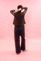 A person viewed from behind wearing a dark brown textured knit vest over sheer black long sleeves and dark navy wide-leg pants, standing against a solid pink studio backdrop.
