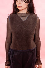 A model wearing a brown textured knit vest over a sheer long-sleeve top and dark pants, set against a solid pink background.