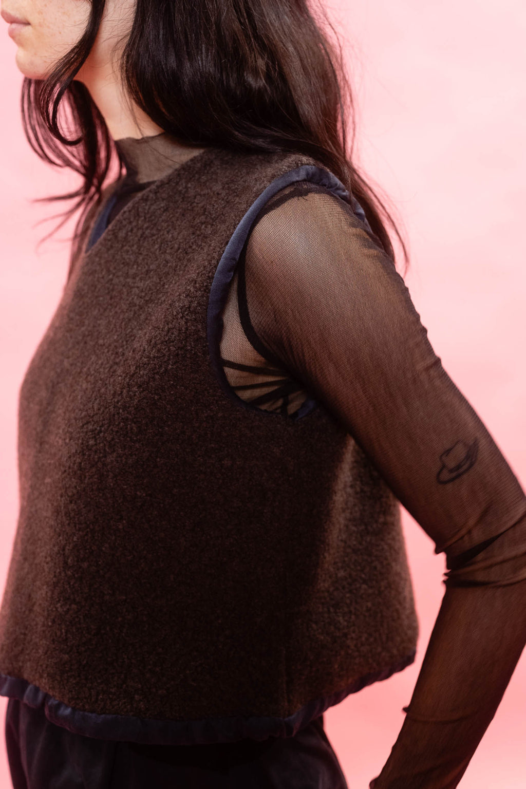 A close-up side view of a model wearing a dark brown textured knit sleeveless vest over sheer dark brown long sleeves, set against a solid pink background.
