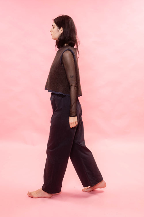 A woman in a dark brown cropped vest, sheer long-sleeve top, and dark wide-leg pants walks barefoot against a solid pink studio backdrop.