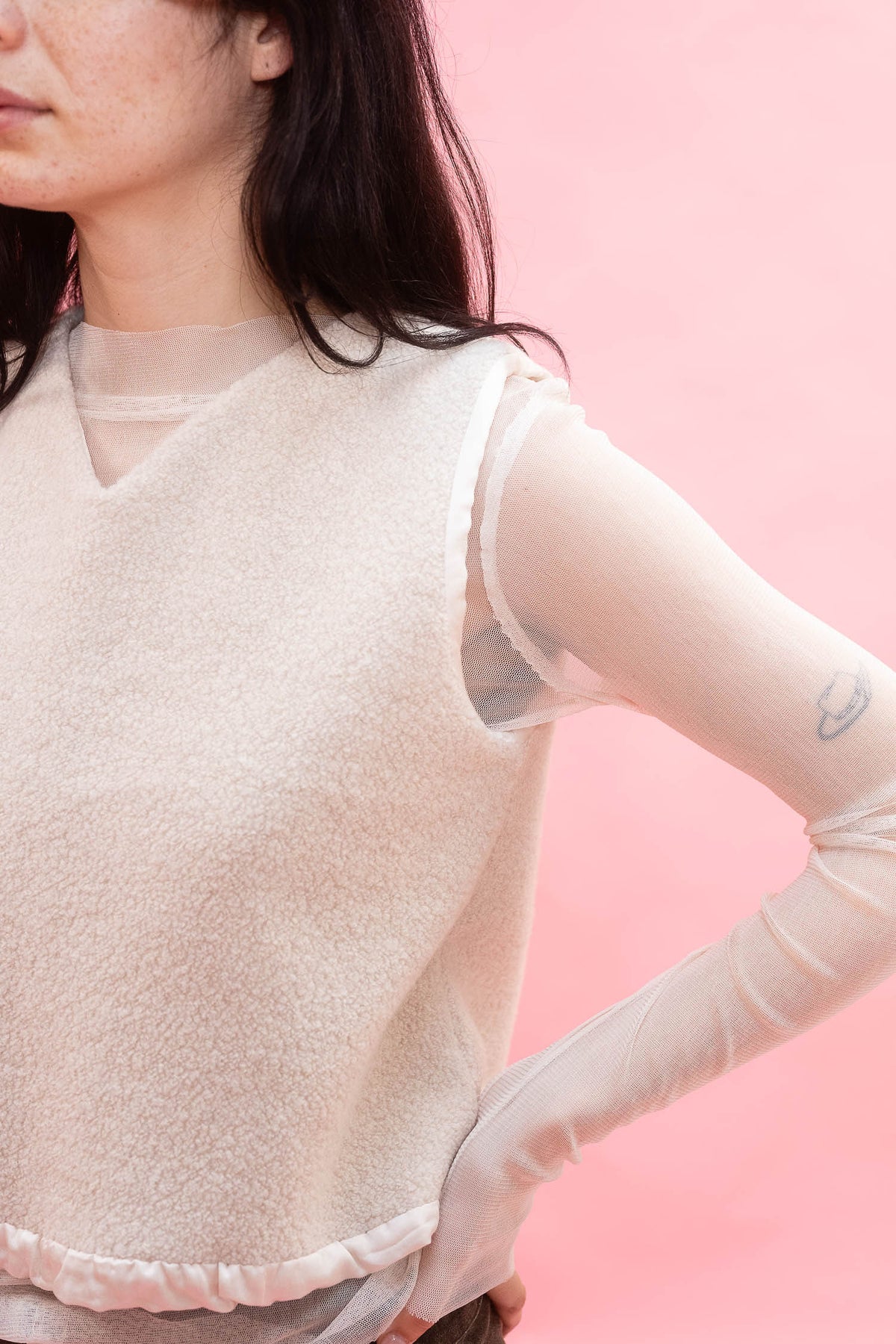 A close-up of a model wearing a cream-colored knit sweater vest over sheer long sleeves, set against a solid pink background.