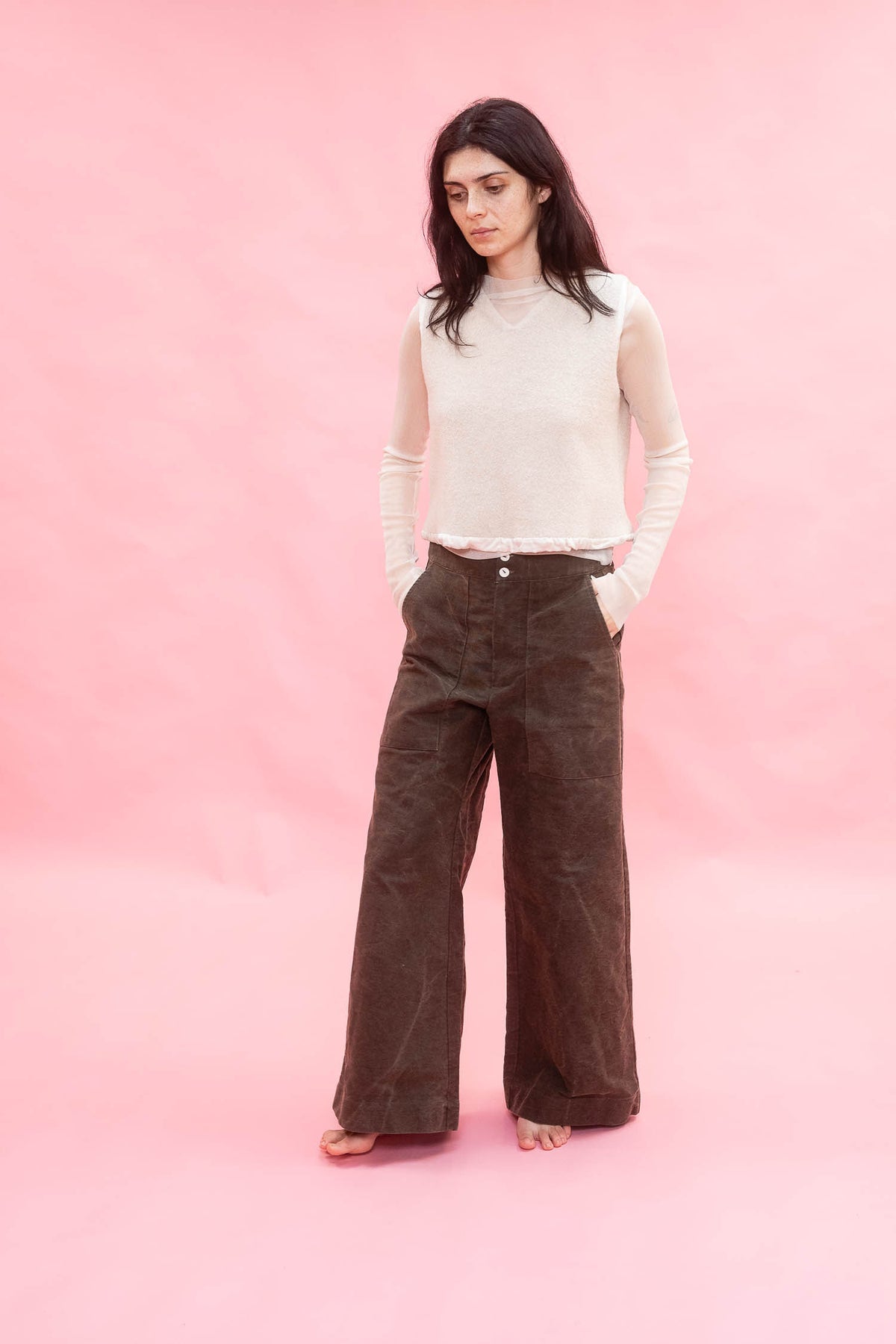 A woman stands against a solid pink studio backdrop wearing a cream ribbed knit long-sleeve top and dark brown wide-leg corduroy pants.