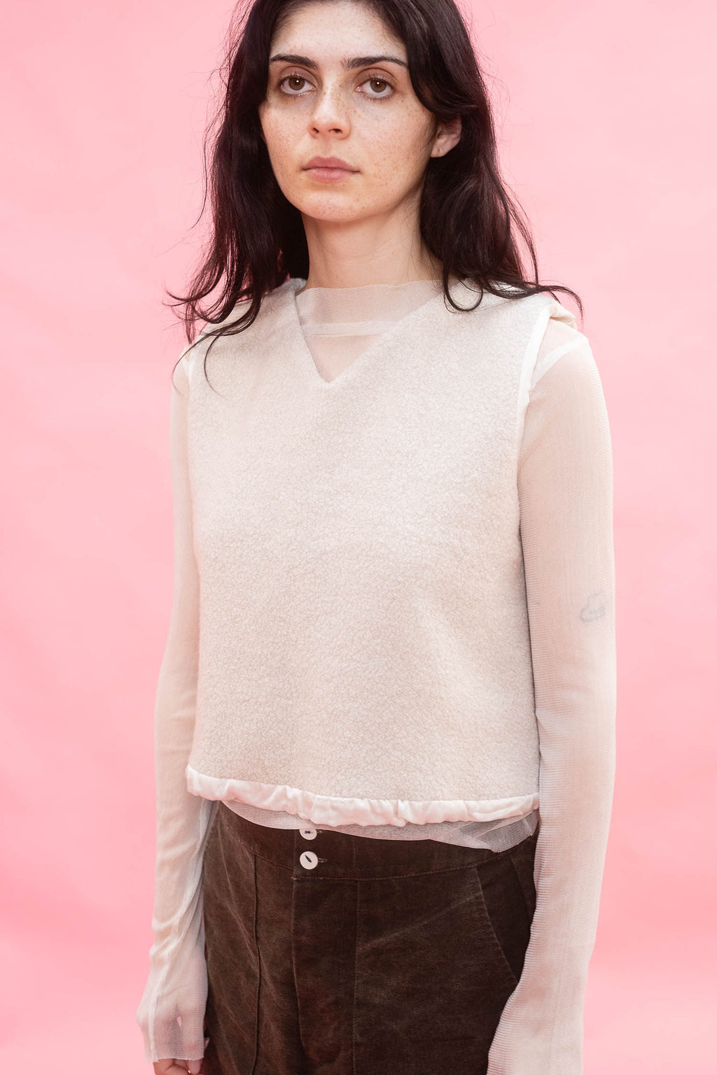 A woman wearing a cream-colored knit sweater vest over a sheer long-sleeve top and brown corduroy trousers, set against a solid pink studio background.