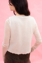 A woman shown from the back wearing a light cream-colored knit top with sheer long sleeves and a structured, slightly cropped back panel, paired with dark brown pants, set against a solid pink background.