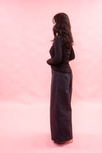 A woman stands in profile against a solid pink studio background, wearing a black long-sleeve top and a very wide, loose navy long pants.