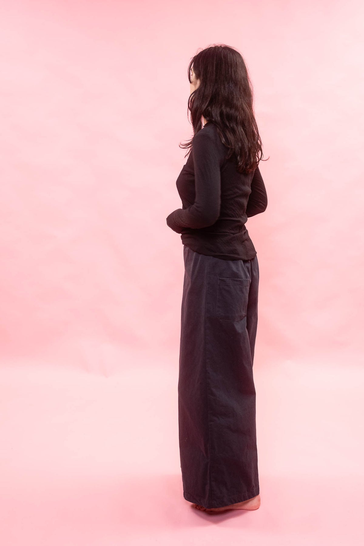 A woman stands in profile against a solid pink studio background, wearing a black long-sleeve top and a very wide, loose navy long pants.