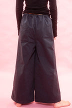A person shown from the back wearing loose, wide-leg dark pants and a black long-sleeve top against a solid pink studio background.