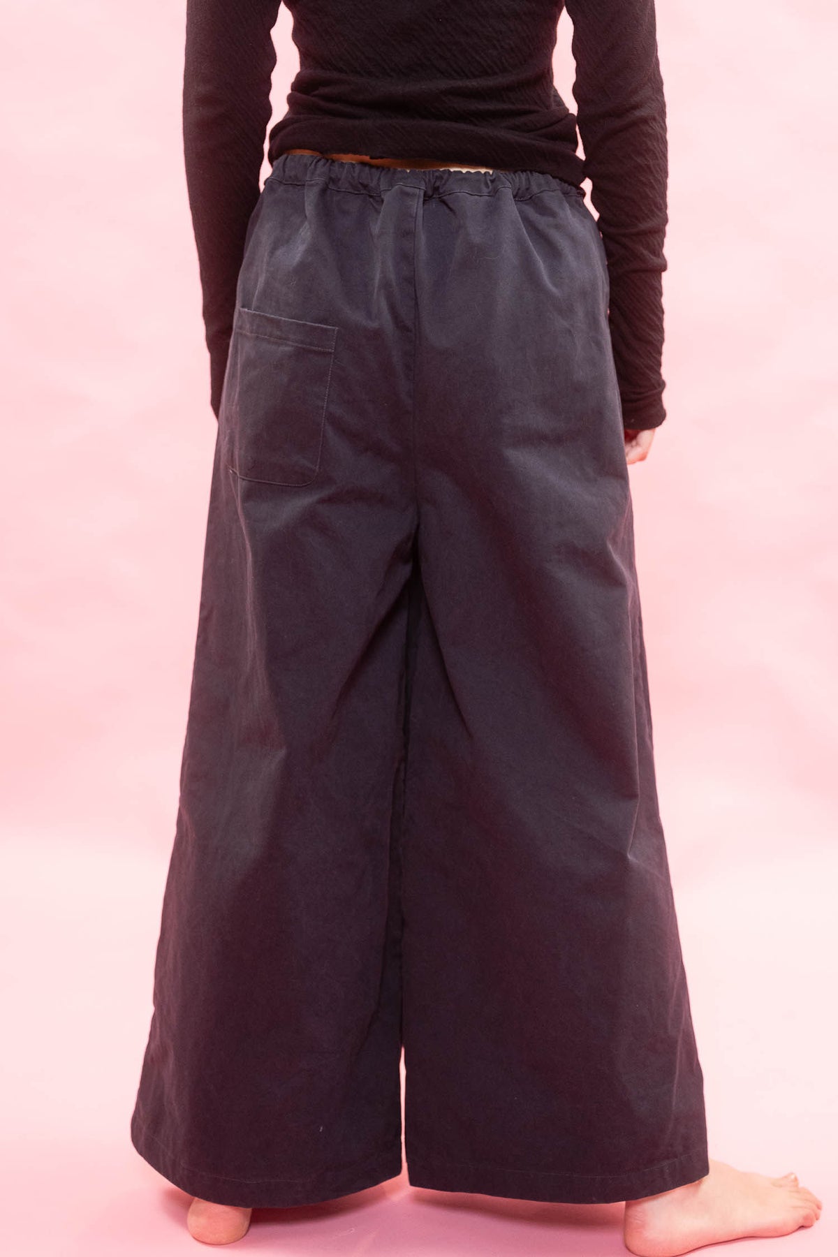 A person shown from the back wearing loose, wide-leg dark pants and a black long-sleeve top against a solid pink studio background.