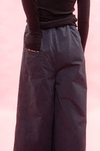 A close-up of a person wearing dark navy wide-leg pants with a side pocket and a black ribbed long-sleeve top, set against a solid pink background.