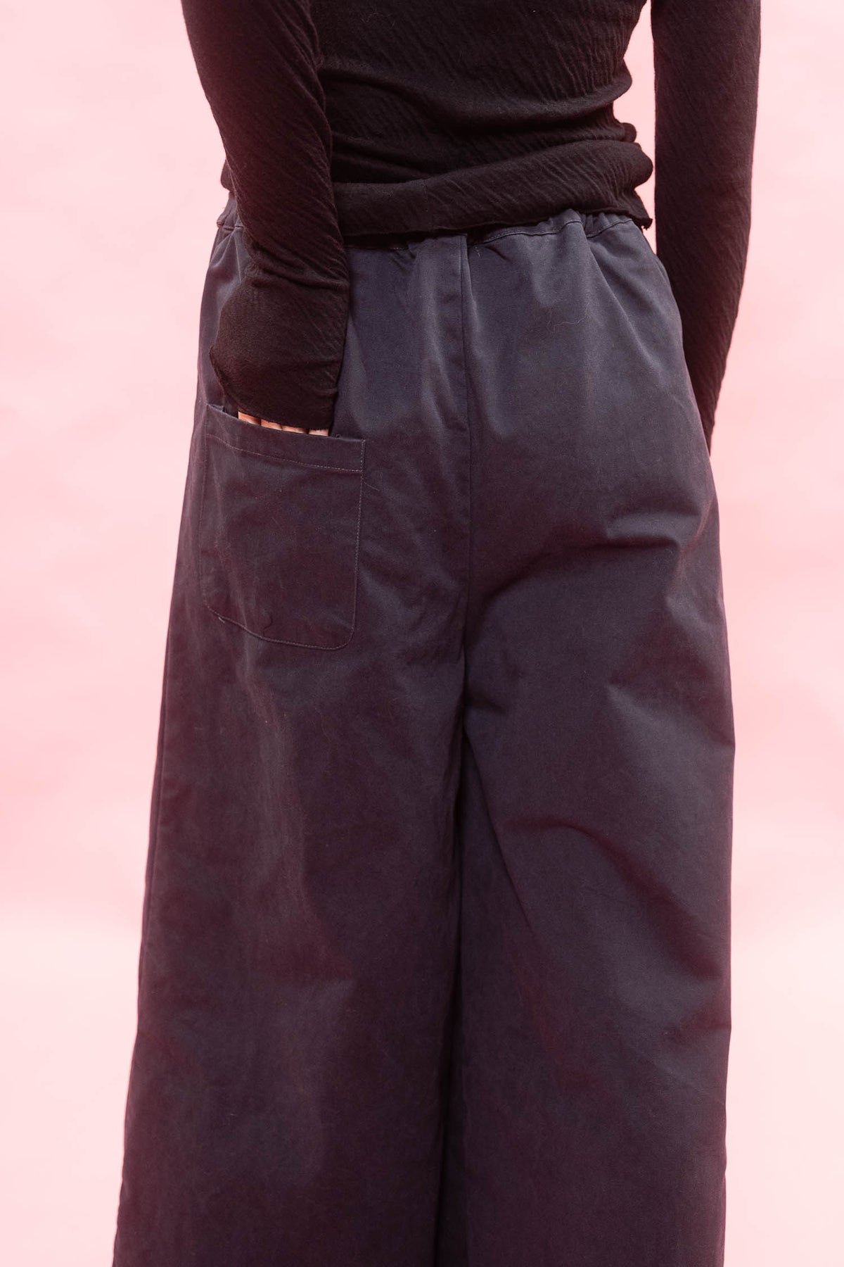 A close-up of a person wearing dark navy wide-leg pants with a side pocket and a black ribbed long-sleeve top, set against a solid pink background.