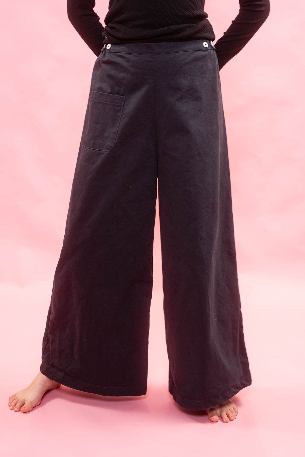 A person wearing very wide-leg navy pants with a black long-sleeve top, standing barefoot against a solid pink studio background.