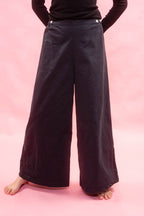 A person wearing very wide-leg navy pants with a black long-sleeve top, standing barefoot against a solid pink studio background.