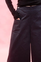 Close-up of a person wearing dark wide-leg trousers with a side pocket and a black long-sleeve top, set against a solid pink background.