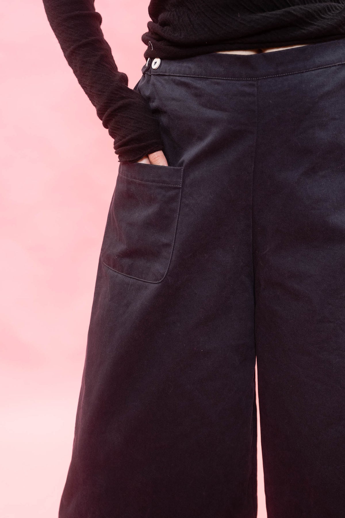 Close-up of a person wearing dark wide-leg trousers with a side pocket and a black long-sleeve top, set against a solid pink background.