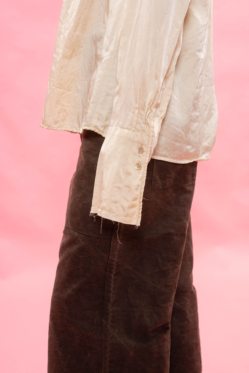 A close-up of a person wearing a cream-colored, satin long-sleeve blouse with raw, frayed edges and a dark brown  pants against a solid pink background.