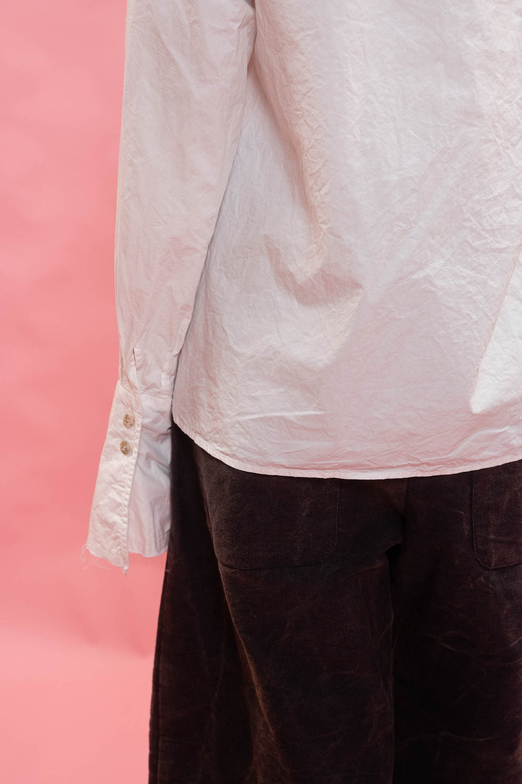 A close-up of a person wearing a white long-sleeve button-up shirt and dark navy cotton trousers, standing against a solid pink background.