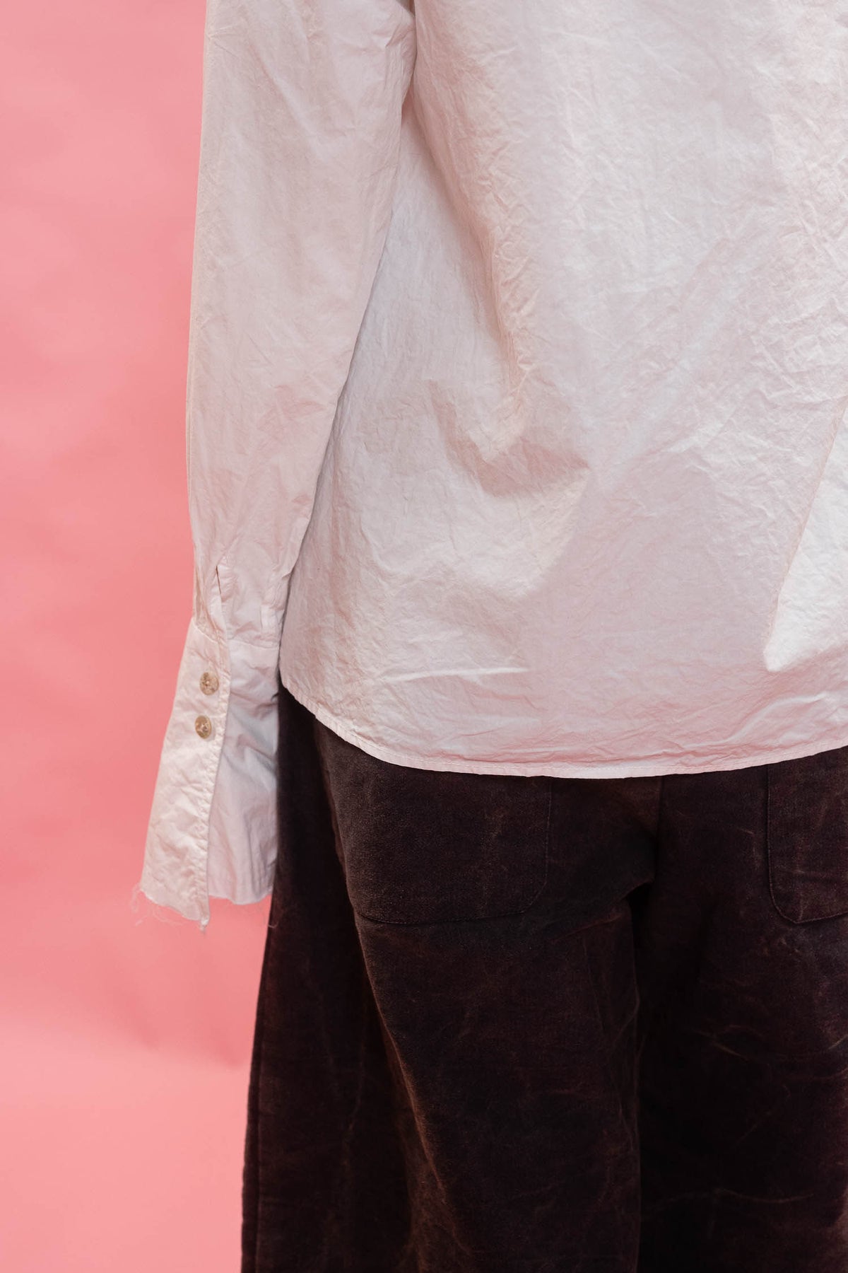 A close-up of a person wearing a white long-sleeve button-up shirt and dark navy cotton trousers, standing against a solid pink background.