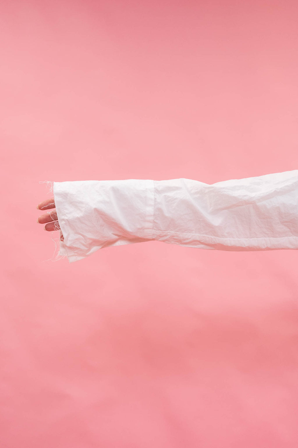 A close-up of a person's hand and forearm wearing a long, oversized white sleeve or cuff, set against a solid pink background.