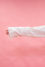A close-up of a person's hand and forearm wearing a long, oversized white sleeve or cuff, set against a solid pink background.