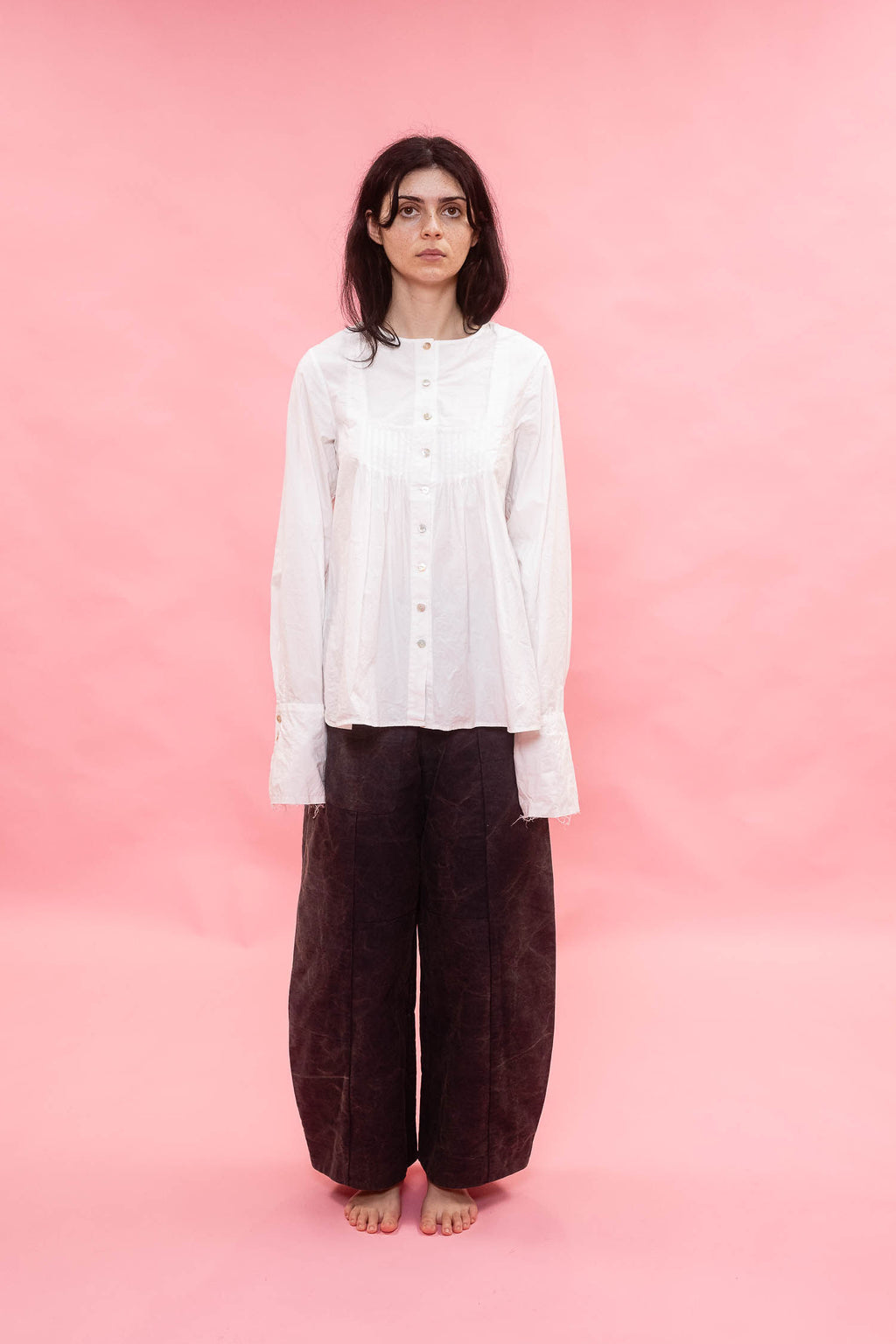 A model stands against a solid pink studio backdrop, wearing a white long-sleeve button-up shirt with ruffled cuffs and dark navy wide-leg trousers.