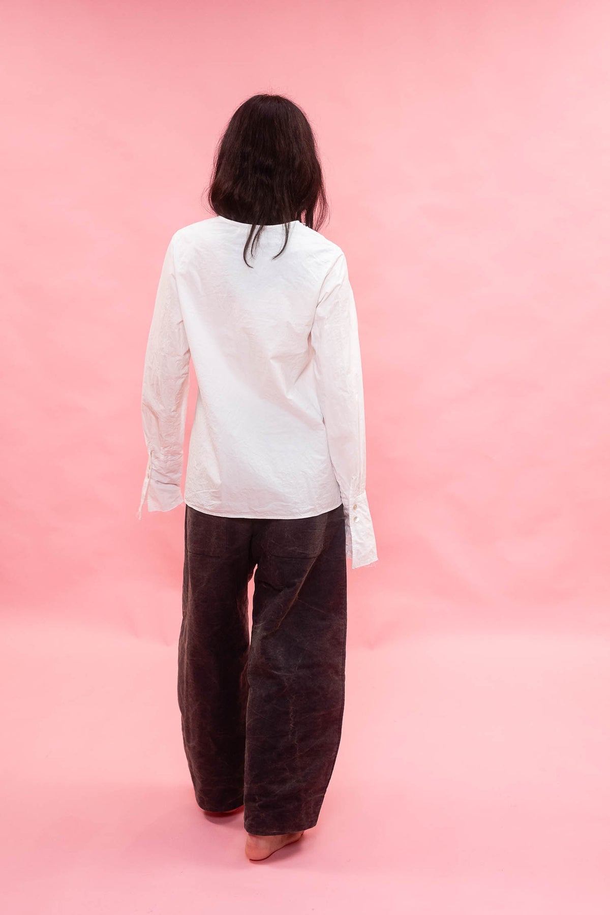 A person shown from the back wearing a white long-sleeve button-up shirt and dark navy wide-leg pants, standing against a solid pink studio backdrop.