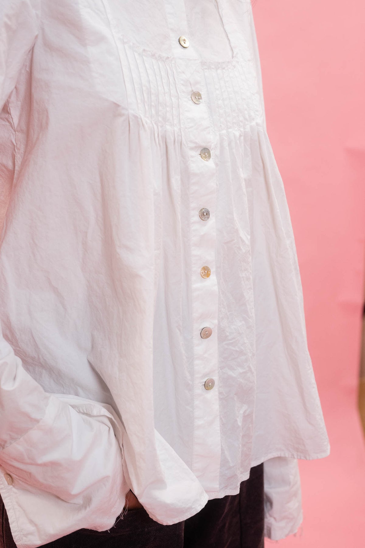 A close-up of a white long-sleeve blouse with a smocked chest panel and a vertical row of mother of pearl buttons, shown against a solid pink background.