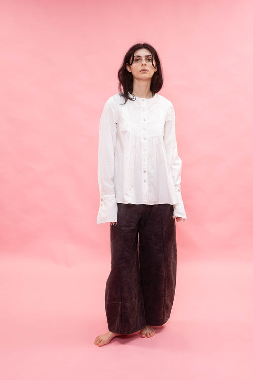 A woman stands barefoot against a solid pink backdrop, wearing a loose white long-sleeve button-up shirt and dark brown wide-leg pants.