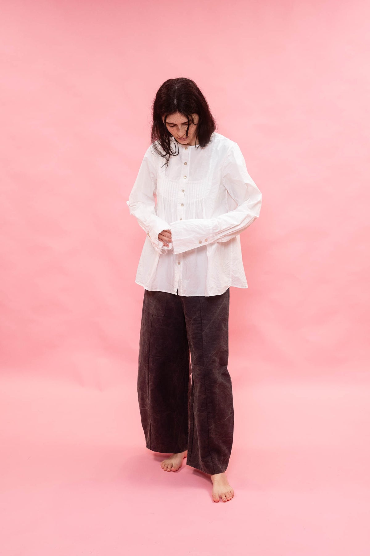 A woman stands barefoot against a solid pink backdrop, wearing a loose white long-sleeve button-up shirt and dark navy wide-leg pants.